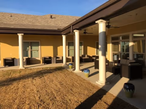 Outdoor covered patio area with several dark wicker chairs and small tables arranged along the wall of a yellow building. The patio is supported by white columns and overlooks a dry grass lawn with decorative ceramic pots placed near the columns.