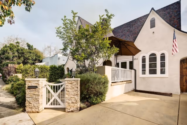Exterior view of a charming residential-style building with a steeply pitched roof and cream-colored walls. The entrance features a small gated area with stone pillars and greenery, including bushes and a tree. An American flag is mounted near the front door, and there is a large umbrella providing shade over a small porch area.
