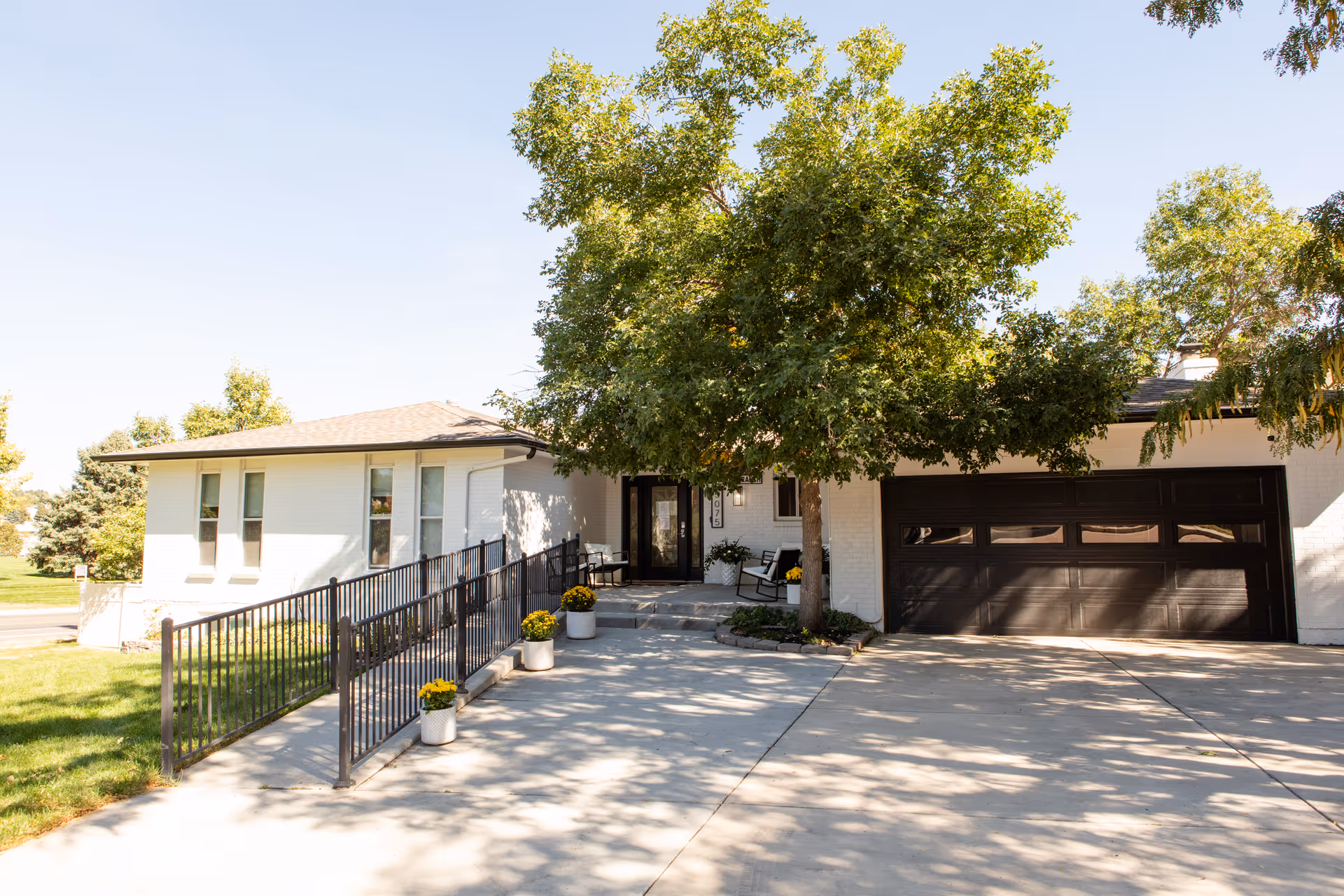 Front exterior view of a single-story white brick building with a dark brown garage door and a concrete driveway. There is a large tree providing shade near the entrance, which has a black door and a ramp with black railings. Yellow flowers in white pots line the ramp and porch area.