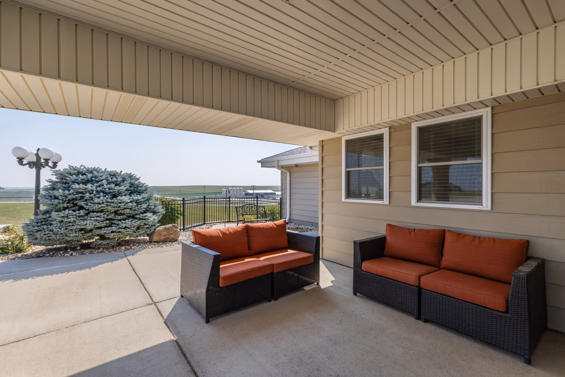 Covered outdoor patio area with two black wicker sofas featuring orange cushions, beige siding walls with two windows, a concrete floor, and a view of a landscaped area with a large bush and a black metal fence under a clear sky.