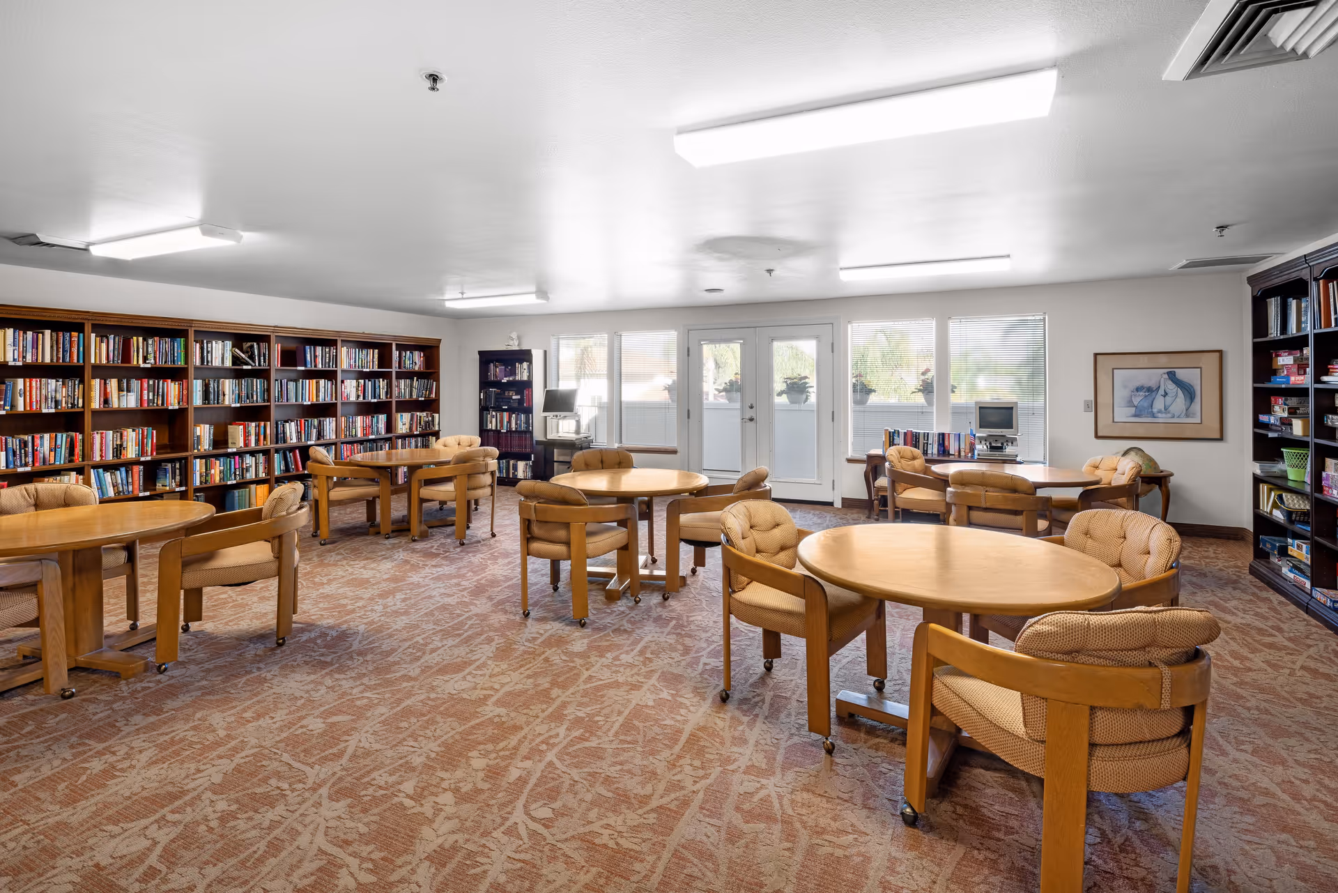 A bright and spacious library or reading room in a senior living community featuring multiple round wooden tables with cushioned chairs on wheels. The room has large windows and glass doors letting in natural light, bookshelves filled with books along the walls, a computer station, and framed artwork on the wall.