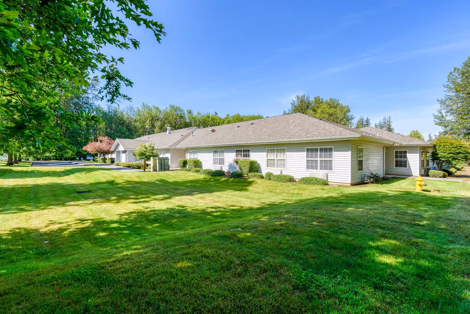 Exterior view of a single-story building with white siding and multiple windows, surrounded by a well-maintained green lawn and trees under a clear blue sky.