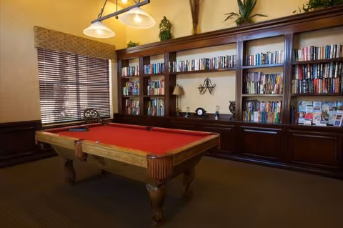 A cozy interior room with a red-felt pool table in front of built-in bookshelves and a window with blinds.
