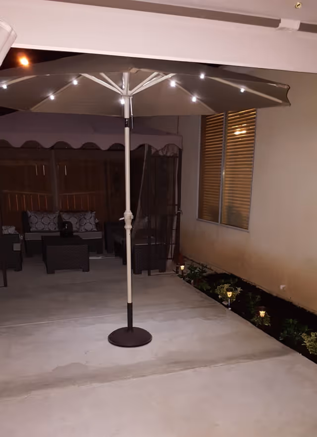 Outdoor patio area at night with a large umbrella featuring small lights around its edge. There is outdoor seating with cushions under a canopy in the background, a window with closed blinds on the right, and small garden lights illuminating plants along the edge of the patio.