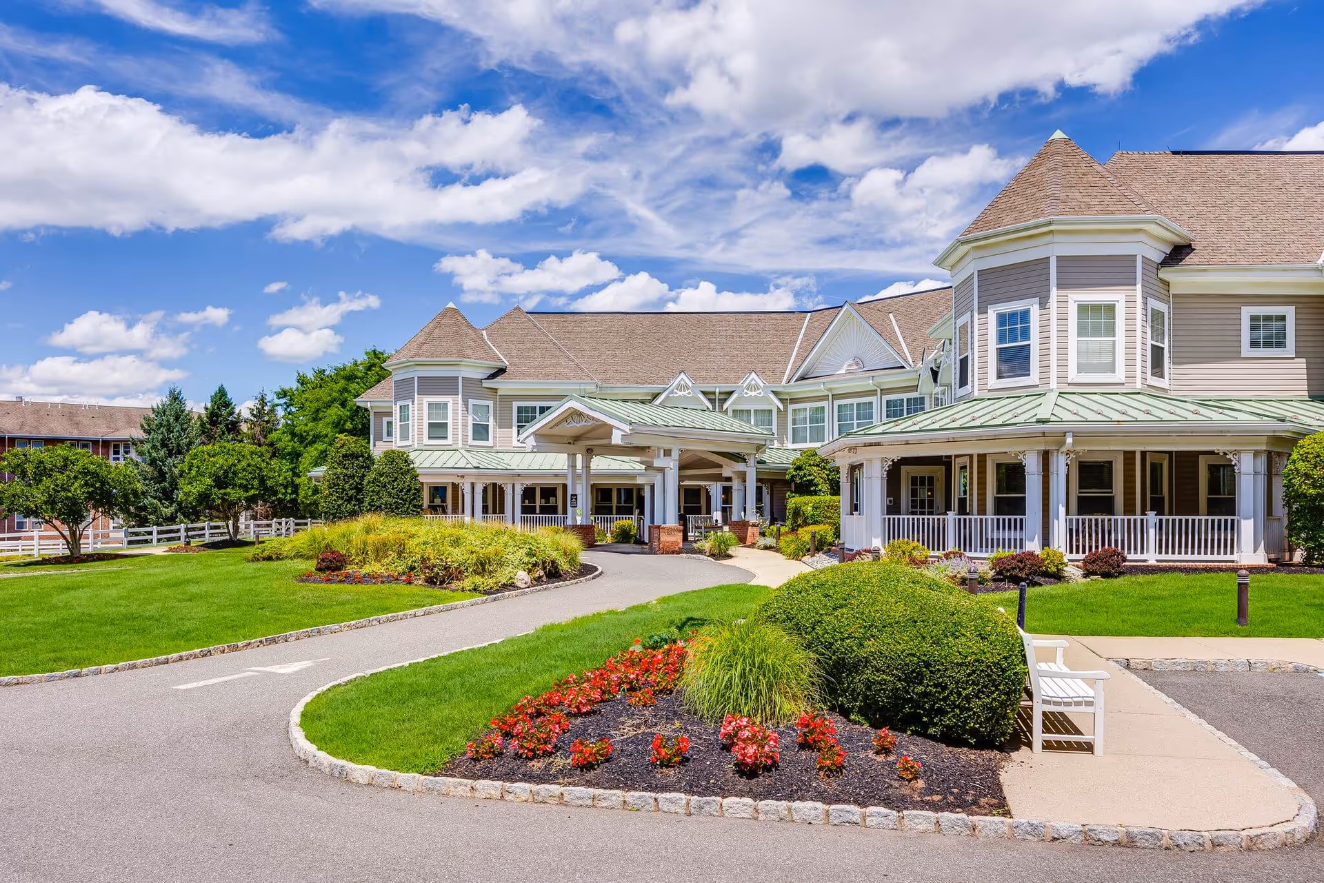 Exterior view of a senior living facility building with a covered entrance, manicured lawns, flower beds, bushes, and a white bench under a partly cloudy blue sky.