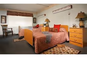 Two twin beds with matching bedding in a well-lit senior facility bedroom with nightstands, lamps, a window, and chairs.
