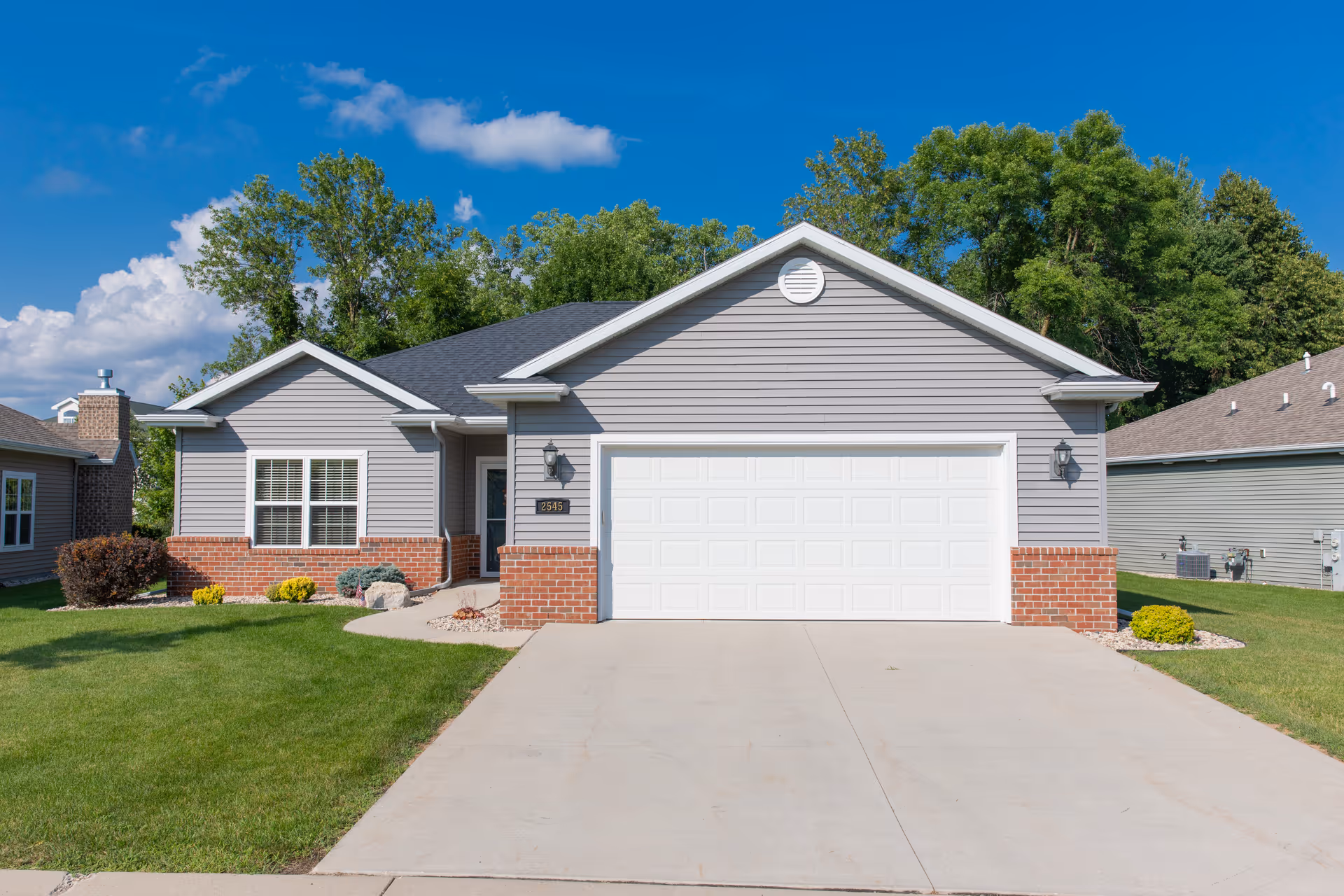 Single-story suburban house with an attached two-car garage, brick accents, and a manicured lawn under a blue sky.