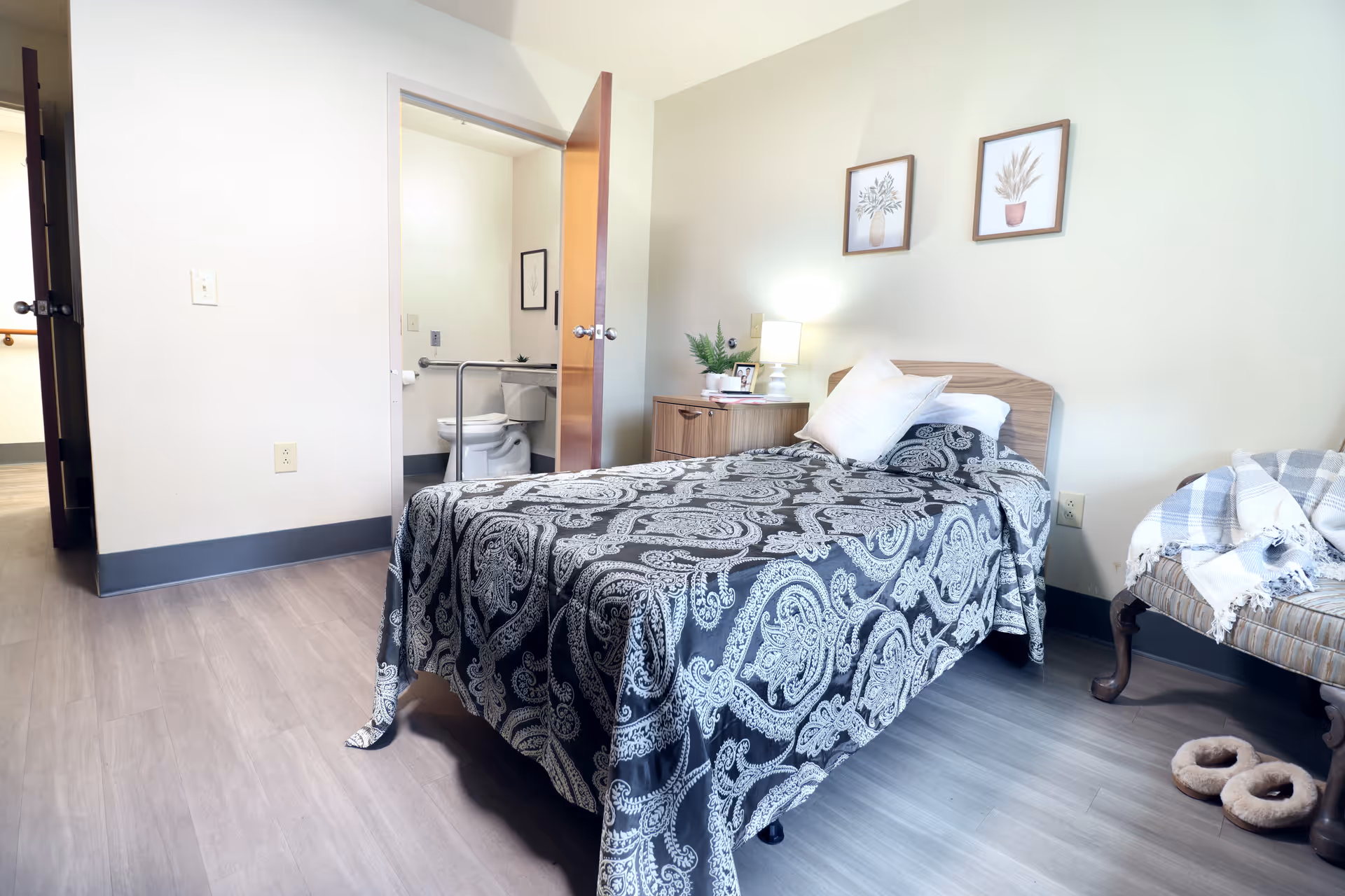 A small bedroom in a senior living facility with a single bed covered in a black and white patterned bedspread. There is a wooden nightstand with a lamp, a plant, and a photo frame next to the bed. Two framed botanical prints hang on the wall above the bed. An open door reveals a bathroom with a toilet and grab bars. A cushioned chair with a plaid blanket and a pair of slippers are on the floor nearby.