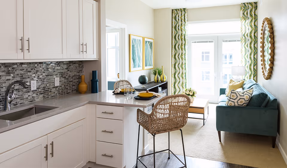 Open-plan kitchen and living area with white cabinets, a breakfast bar with a wicker stool, and a teal sofa by a sunlit window.