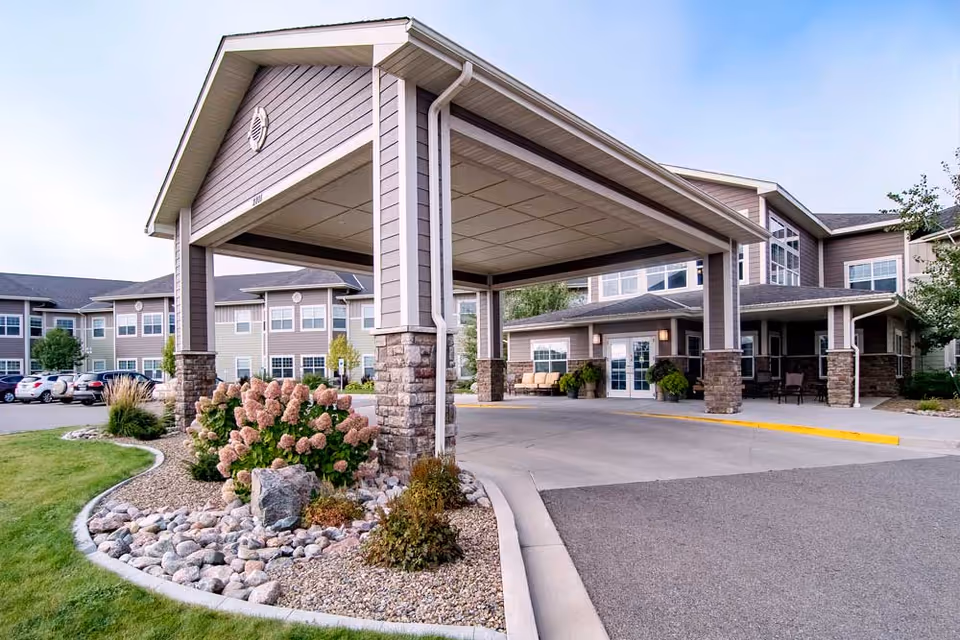 Entrance of a senior living facility with a covered drop-off area supported by stone pillars, landscaped garden with bushes and rocks, and a multi-story building with many windows in the background.