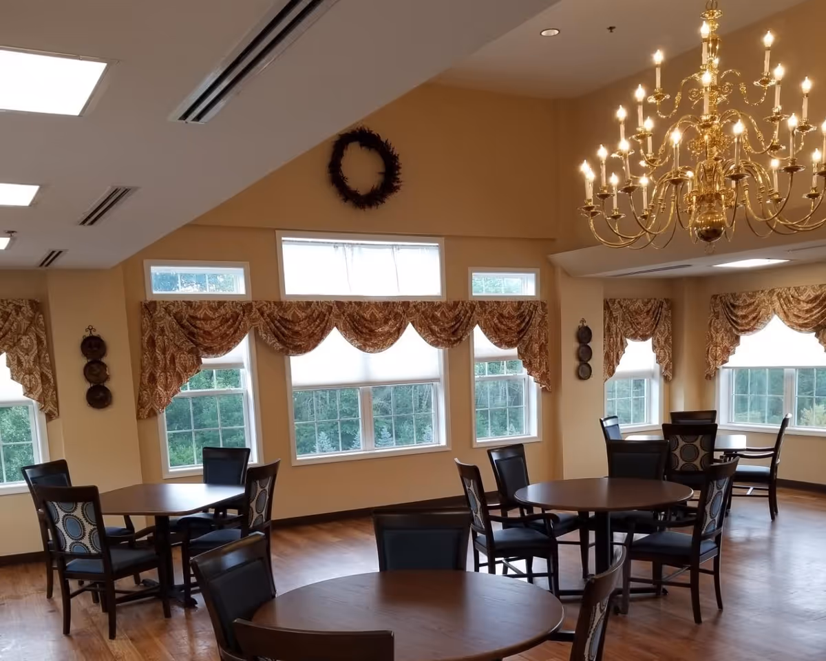 Dining room with round wooden tables and chairs, large windows with patterned valances, and a brass chandelier.