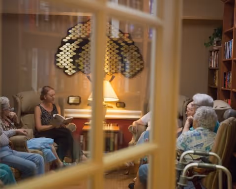 A group of elderly residents seated in a cozy common room listening to a woman reading aloud.