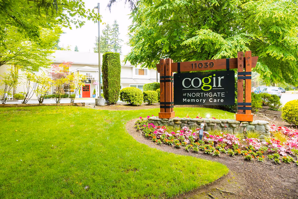 Outdoor view of Cogir of Northgate Memory Care facility with a large wooden sign displaying the facility name and address 11039, surrounded by green grass, colorful flowers, and trees. The building entrance with a red door is visible in the background.