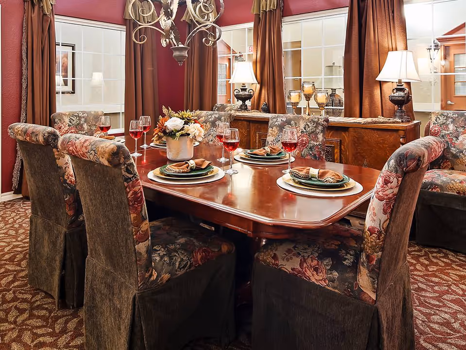 Elegant dining room with a polished wooden table set for four, floral-upholstered chairs, decorative centerpiece and warm lamps.