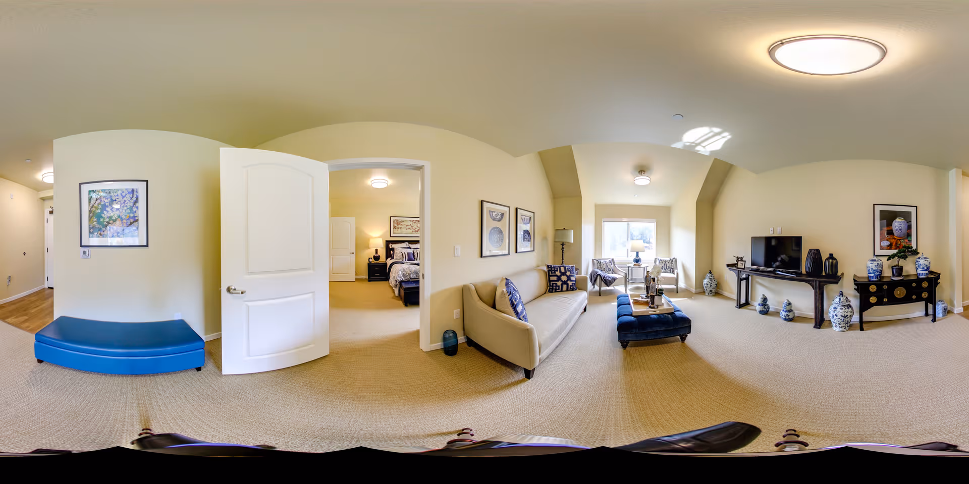 Spacious furnished living room with a sofa, ottoman, TV console, decorative vases and a doorway leading to a bedroom.