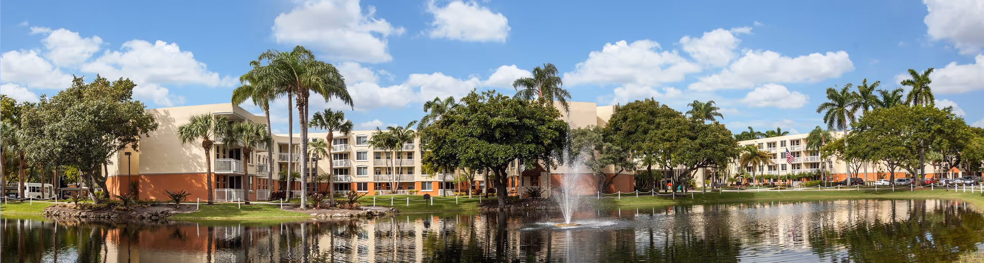 Exterior view of Five Star Premier Residences of Hollywood showing a multi-story building surrounded by palm trees and other greenery, with a pond and a water fountain in the foreground under a partly cloudy blue sky.