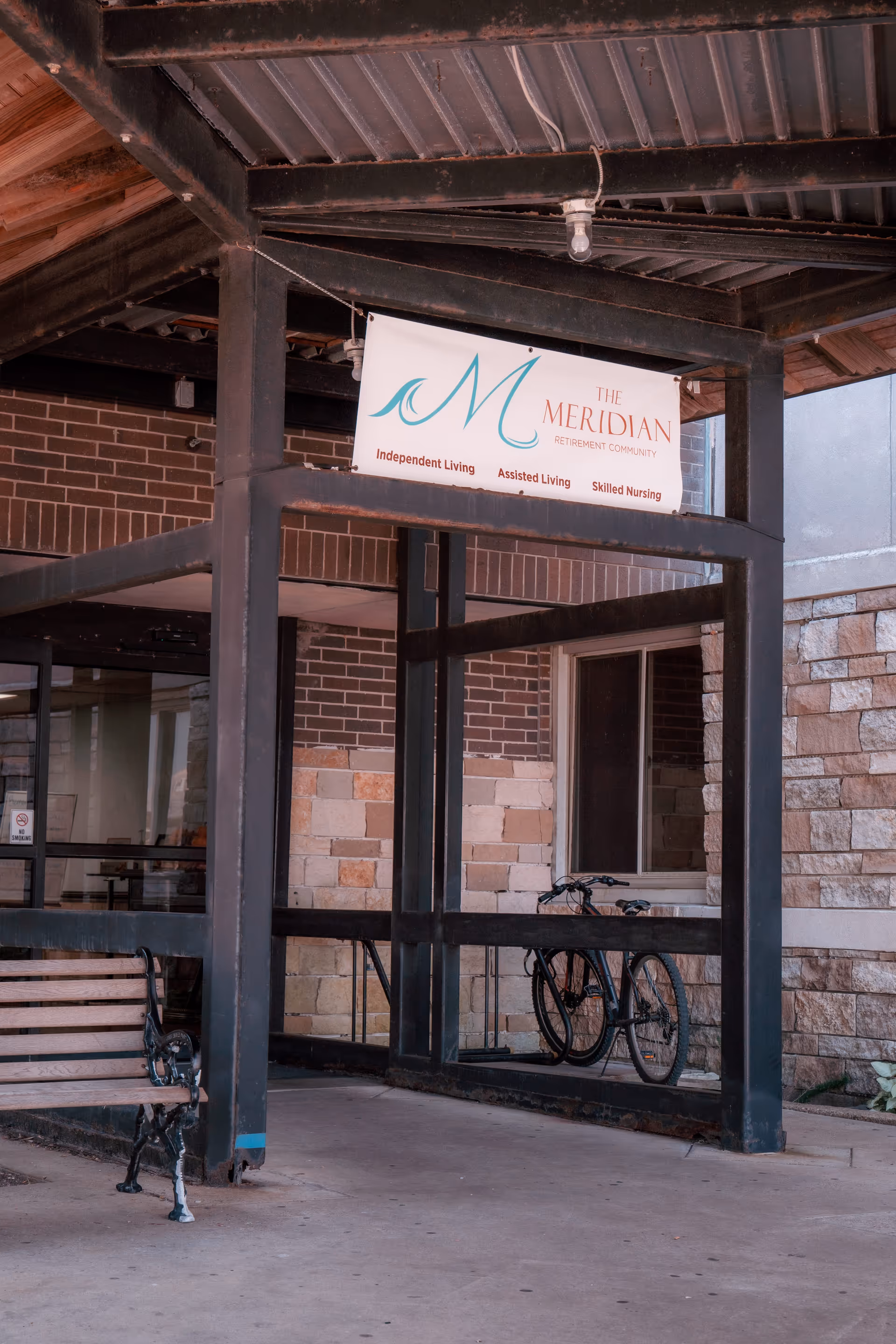 Covered entrance of The Meridian retirement community showing a sign, a bench, and a parked bicycle.