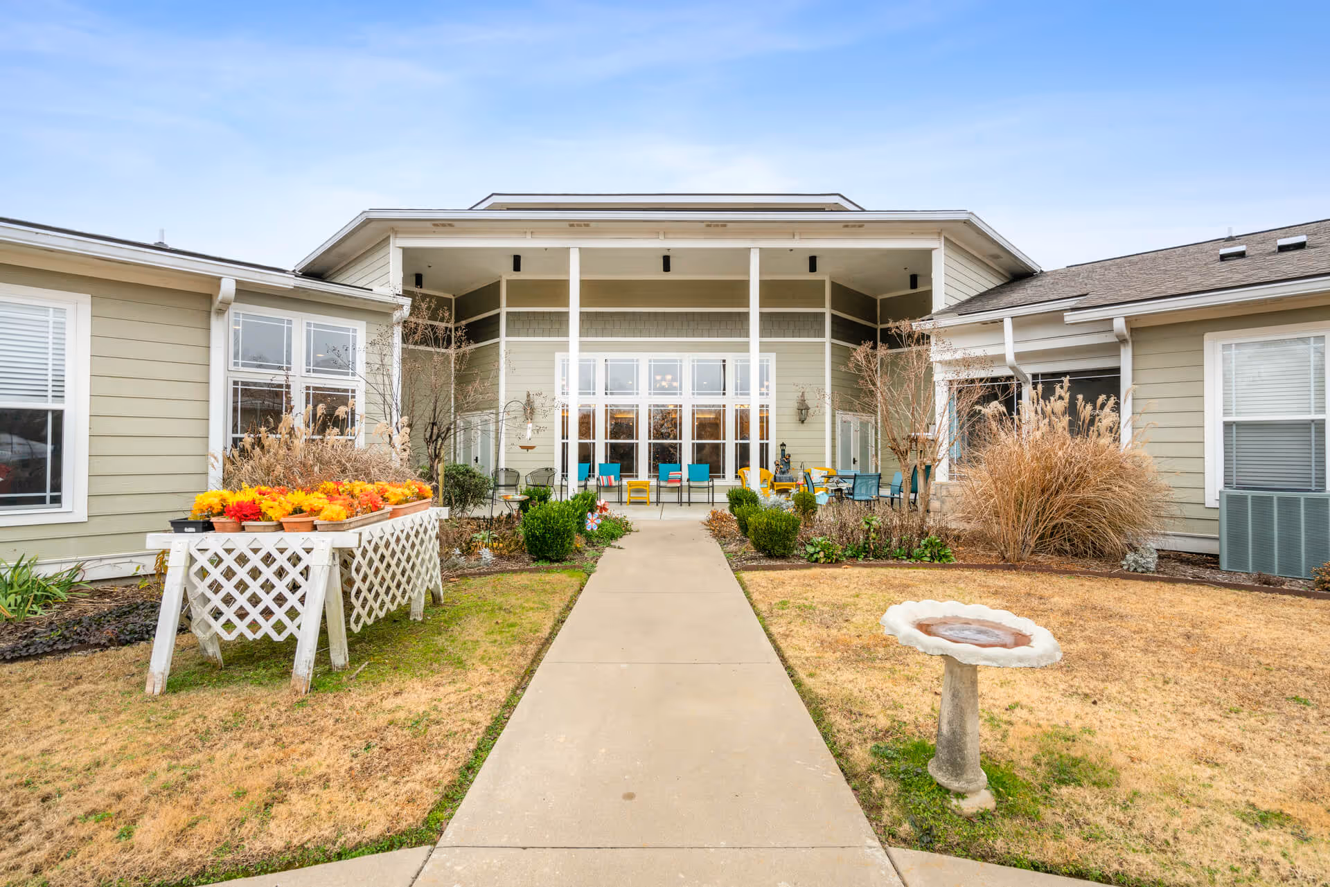 Exterior view of StoneBridge Senior Living - Conway showing a paved walkway leading to a covered patio area with colorful chairs. The building is light green with white trim, surrounded by landscaped bushes, ornamental grasses, a birdbath, and a raised flower bed with bright yellow and orange flowers.
