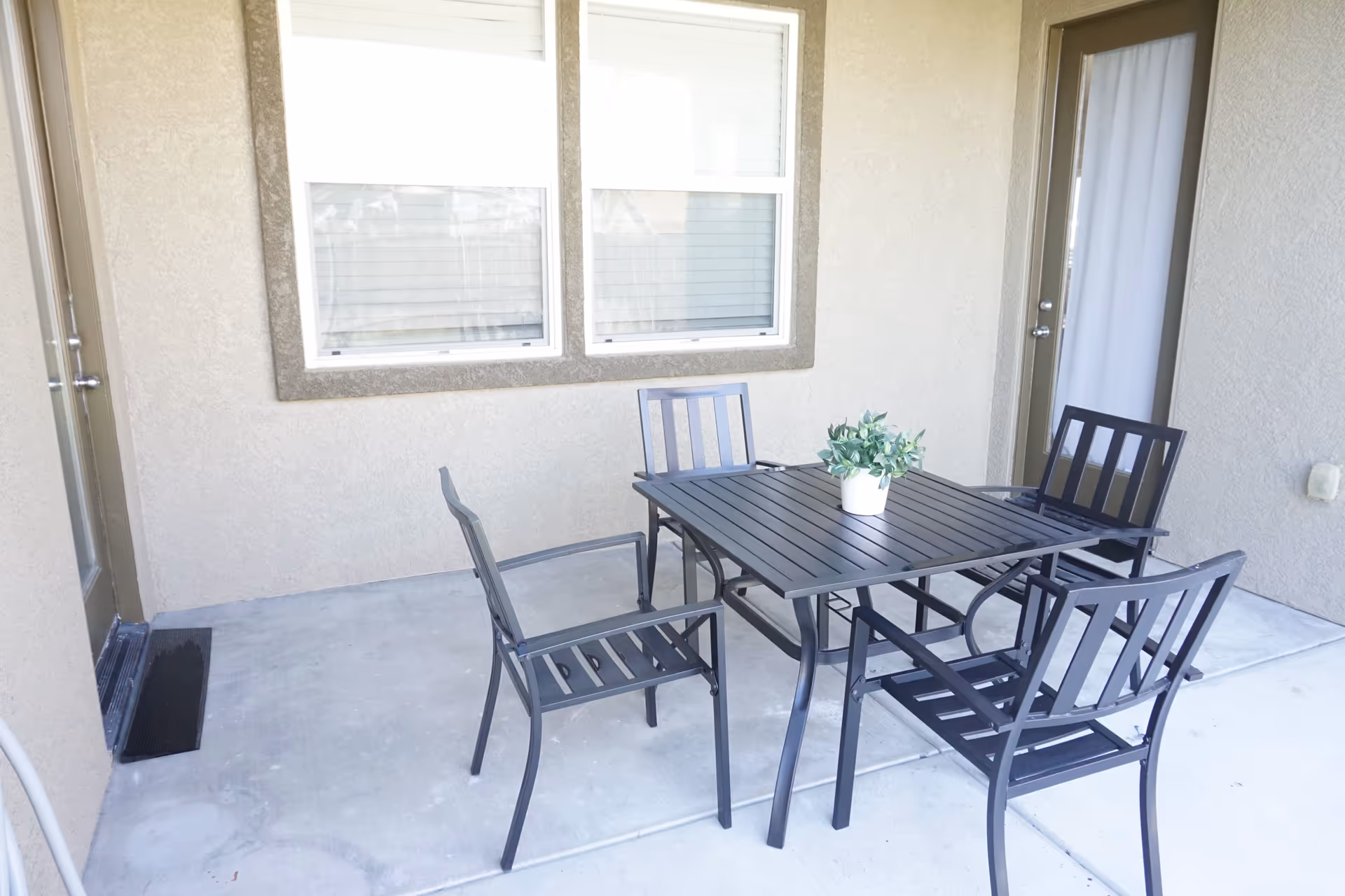 Covered patio with a black metal dining table, four chairs, and a small potted plant in front of windows and doors.