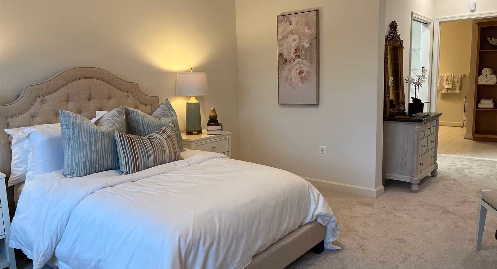 A neatly made bed with a beige upholstered headboard and multiple decorative pillows in shades of blue and striped patterns. A nightstand with a lamp and decorative items is beside the bed. The room has beige walls and carpeted floor. A dresser with a mirror and a potted orchid is visible near an open doorway leading to a bathroom with towels and shelves.