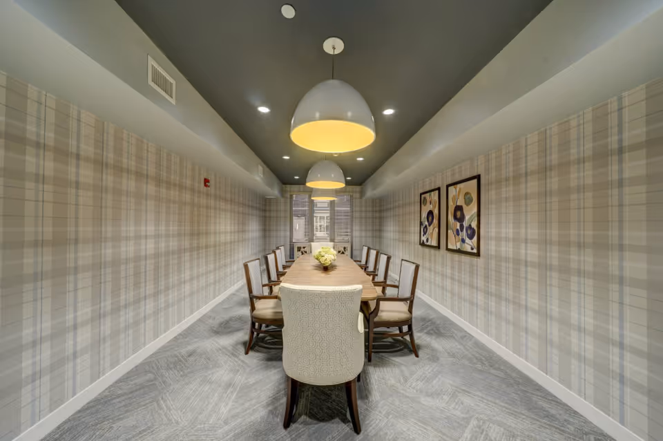 A modern dining room with a long wooden table surrounded by ten chairs, featuring patterned wallpaper with a plaid design, two framed floral artworks on the right wall, and three large pendant lights hanging from a dark ceiling.