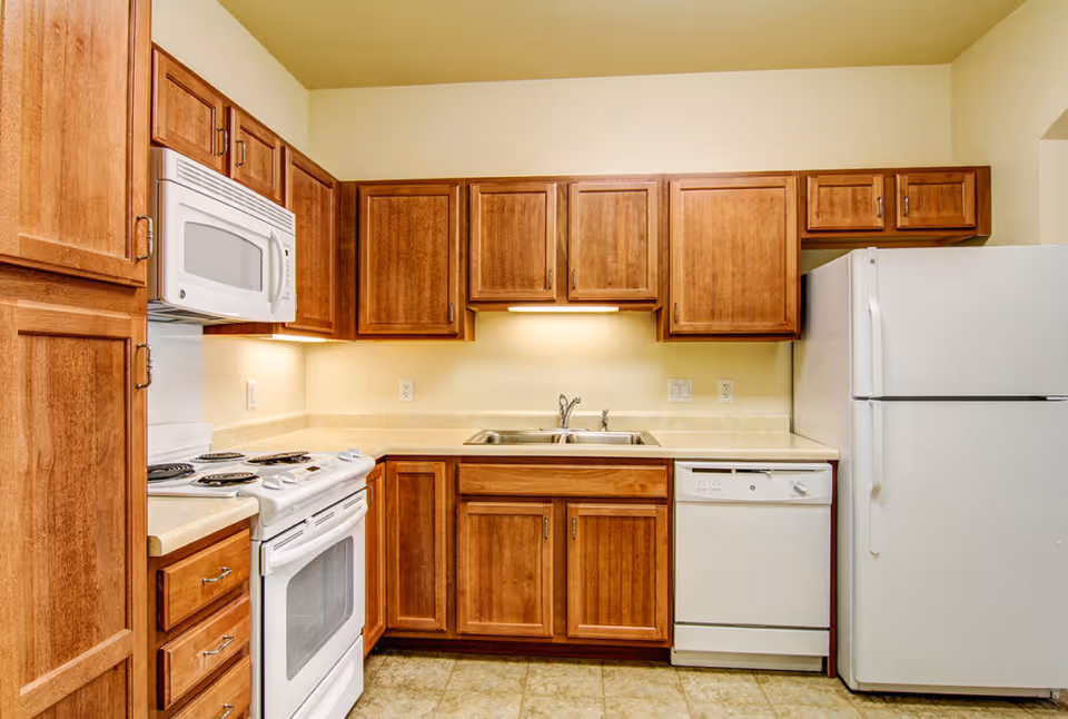 A kitchen with wooden cabinets, a white electric stove with oven, a white microwave above the stove, a double sink with a faucet, a white dishwasher, and a white refrigerator. The countertops are light-colored, and the floor has beige tiles.