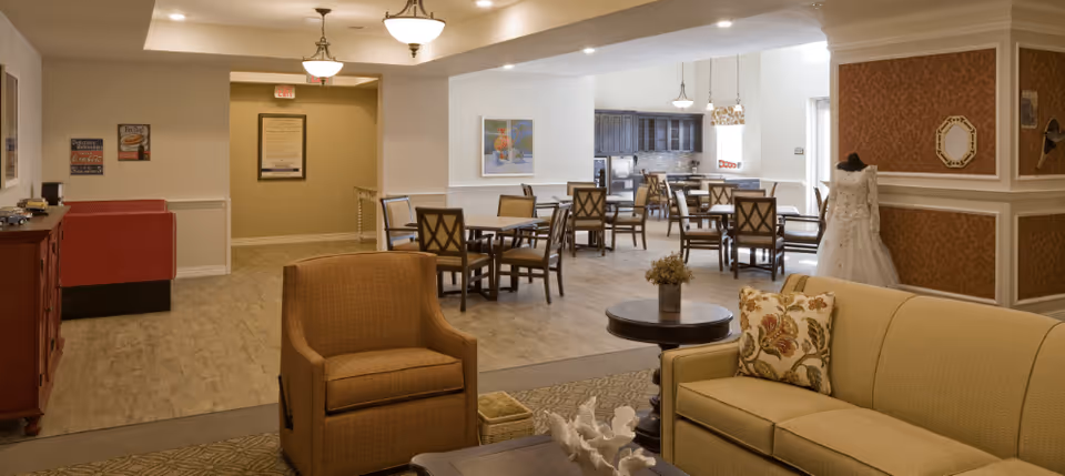 A spacious senior living facility common area featuring a beige sofa with floral pillows, a brown armchair, and a round coffee table with a small plant. In the background, there are multiple dining tables with chairs, a kitchen area with dark wooden cabinets, and a mannequin dressed in a white gown. The walls are decorated with framed pictures and patterned wallpaper, and the floor is a combination of carpet and wood.