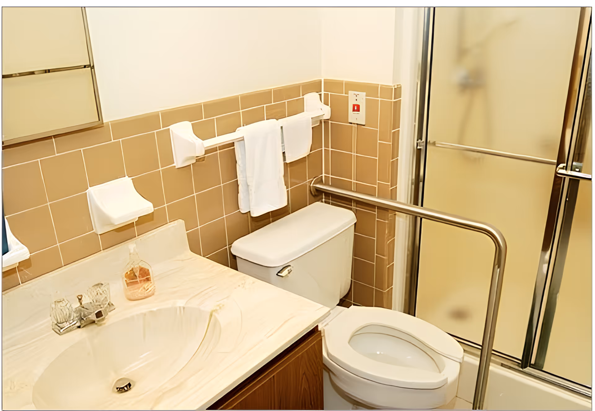 A bathroom with beige tiled walls, a white sink with a marble countertop, a toilet with a raised seat, a metal grab bar beside the toilet, a towel rack with white towels, and a shower with frosted glass sliding doors.
