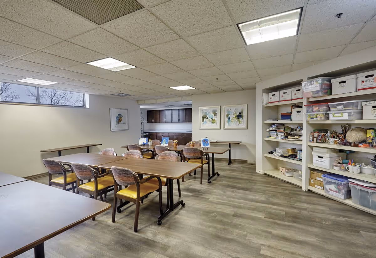 A well-lit activity room in a senior living facility with several tables and chairs arranged for group activities. Shelves on the right side hold various craft supplies and storage boxes. The room has light-colored walls, wood flooring, and ceiling lights. Artwork is displayed on the walls, and a window near the ceiling lets in natural light.
