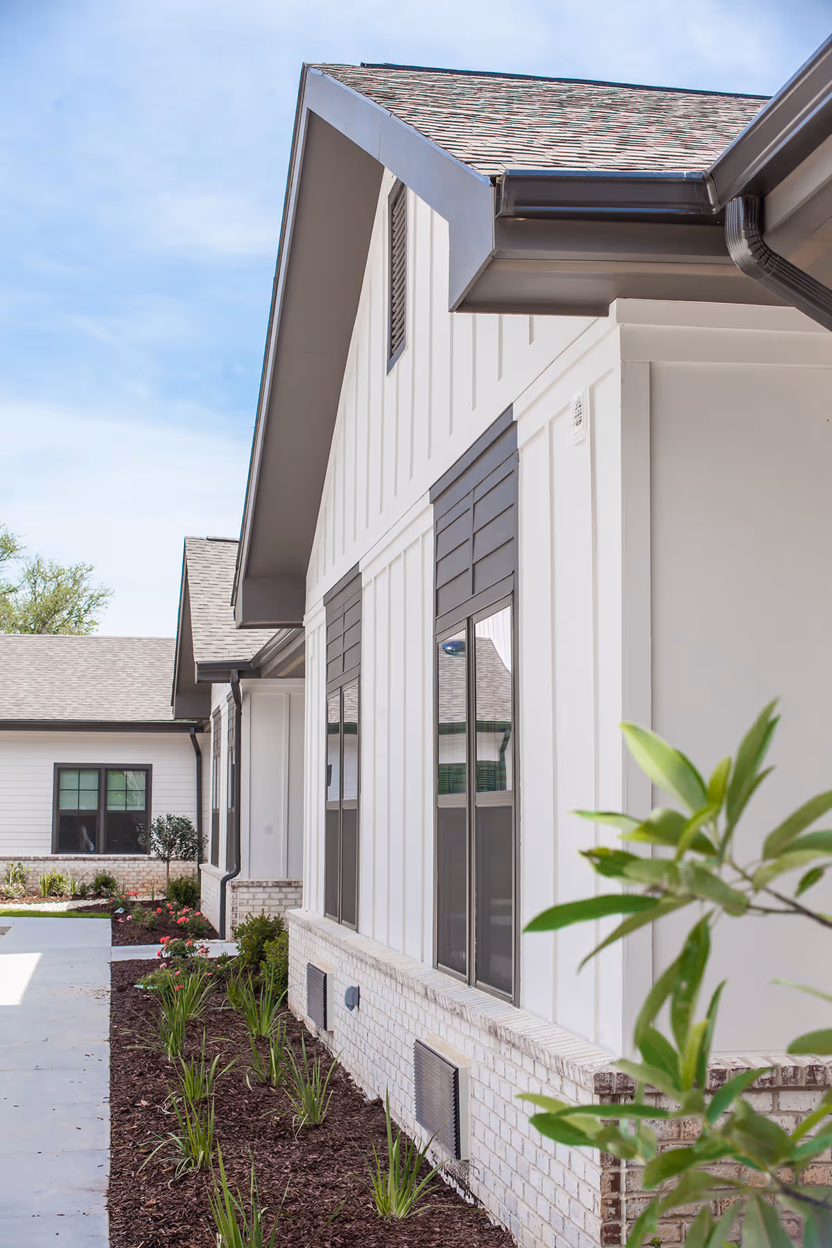 Exterior view of a modern building with white siding and brick foundation, featuring multiple windows and a landscaped garden with green plants and mulch along the walkway under a clear blue sky.