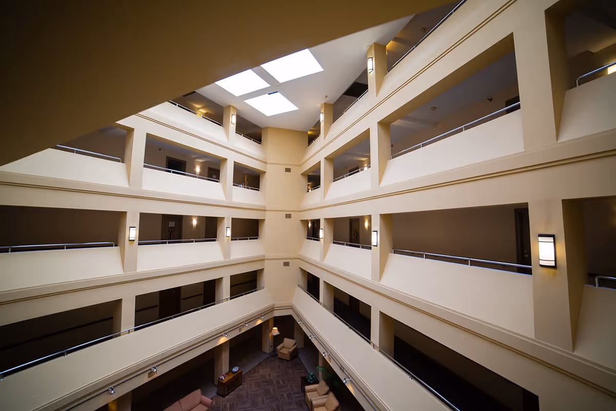 Interior view of a multi-story atrium in a senior living facility with beige walls, railings along each floor, ceiling skylights, and seating areas on the ground floor.