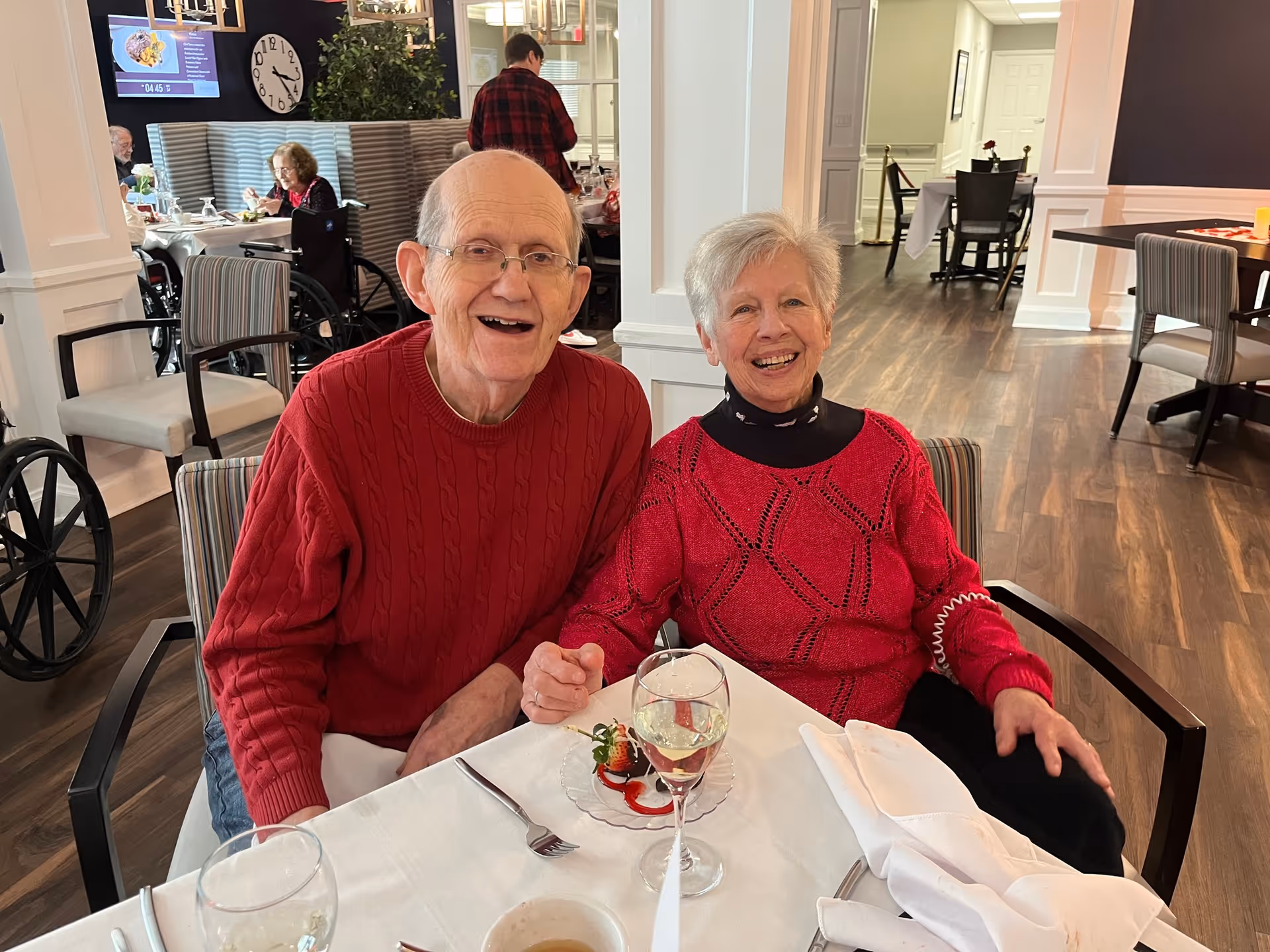 An elderly man and woman sitting together at a dining table in a senior living facility, both smiling and wearing red sweaters. The table is set with a glass of white wine, a dessert plate with strawberries, and white napkins. In the background, other residents are seated and dining in a well-lit room with wooden floors and striped chairs.