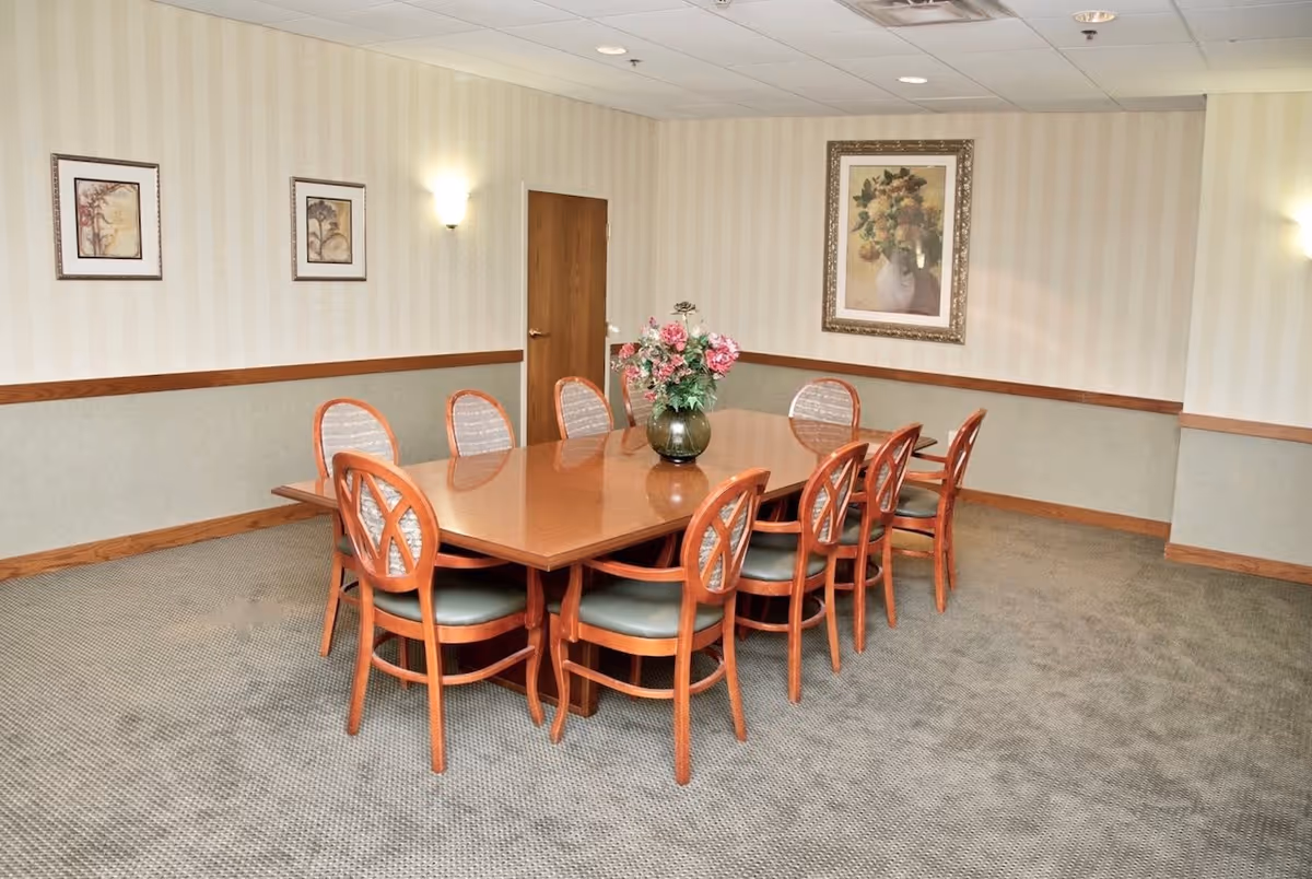 A dining room with a rectangular wooden table surrounded by ten wooden chairs with cushioned seats. A vase with pink and white flowers is placed in the center of the table. The room has beige wallpaper with a subtle pattern, three framed floral paintings on the walls, and wall-mounted lights. The floor is carpeted in a neutral tone.