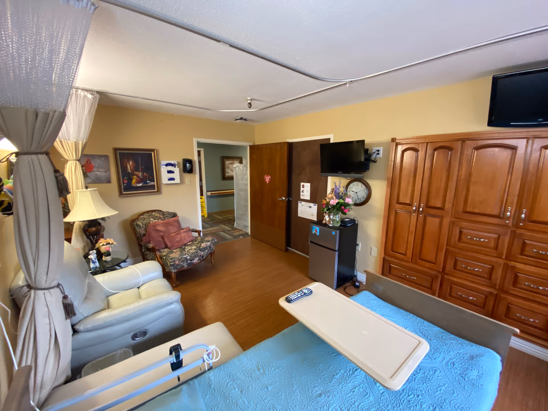 A furnished senior care bedroom with a bed and overbed table, recliner and armchair, TV, wooden cabinets, and a doorway to a hallway.