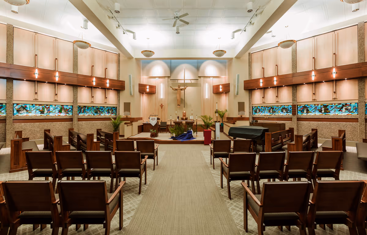 Interior view of a chapel with wooden pews arranged in rows facing an altar. The altar features a crucifix mounted on the wall, candles, and religious decorations. The walls have stained glass windows with colorful designs, and the ceiling has recessed lighting and ceiling fans.