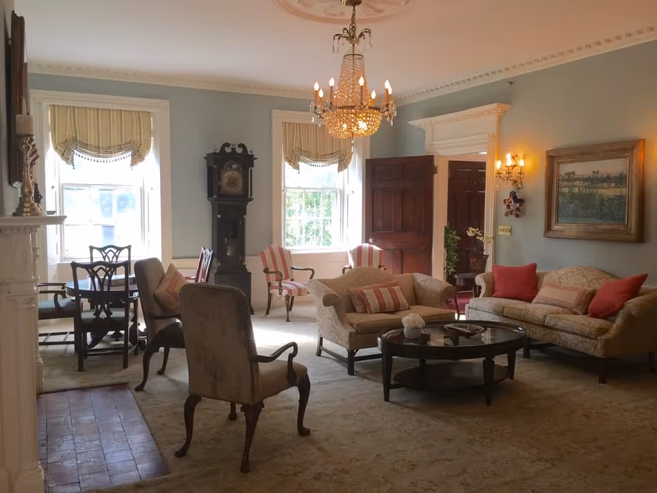 A well-lit living room with classic decor featuring a crystal chandelier, two large windows with striped valances, a grandfather clock, a fireplace, and a seating area with two sofas, armchairs, and a round glass coffee table. The walls are painted light blue and adorned with a framed landscape painting and wall sconces.