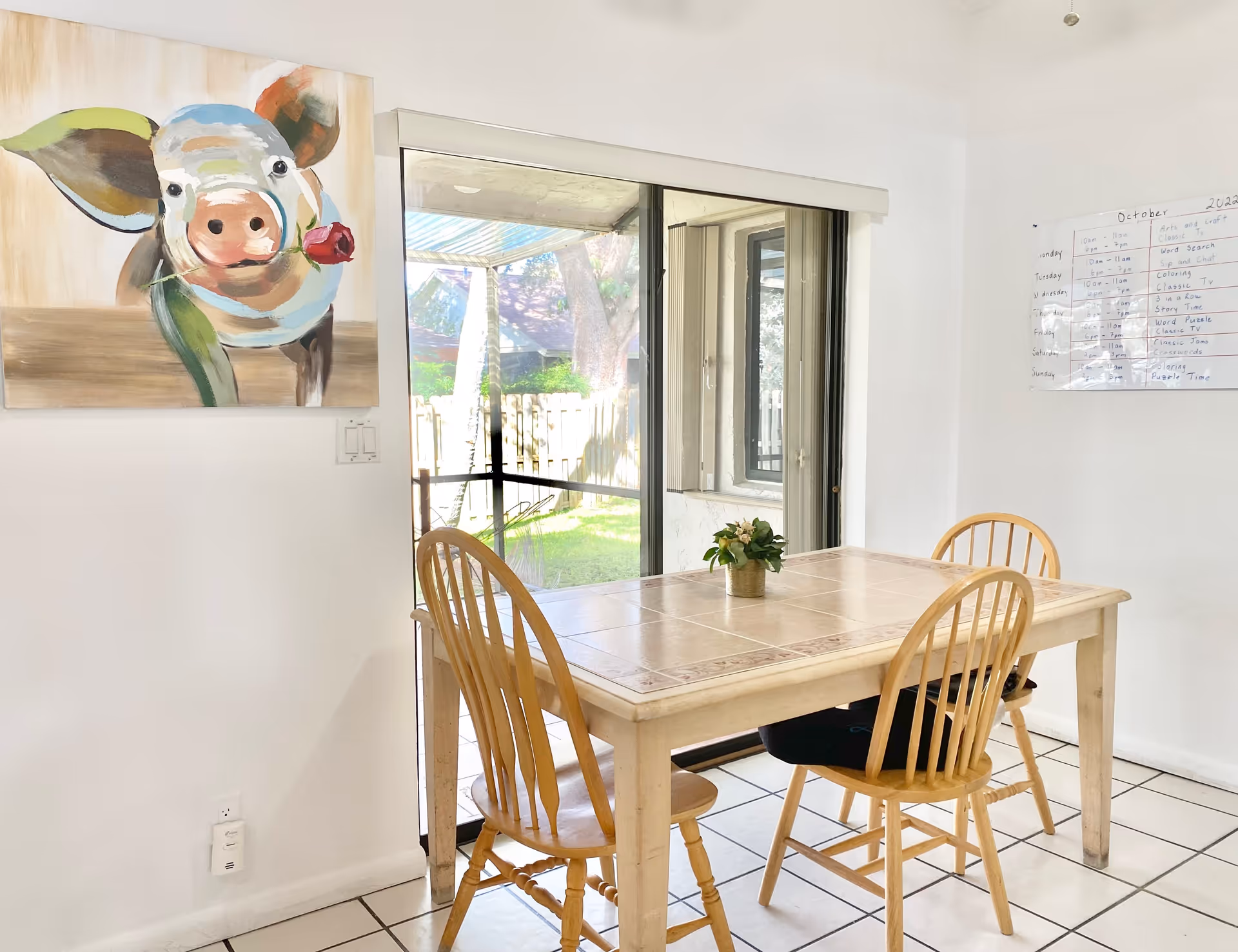 A bright dining area with a wooden table and four matching wooden chairs. A small potted plant is placed in the center of the table. Behind the table is a large sliding glass door leading to a fenced backyard with green grass and trees. On the left wall, there is a colorful painting of a pig holding a rose in its mouth. On the right wall, a whiteboard with a weekly schedule is visible.