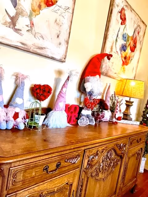 A wooden sideboard with intricate carvings is decorated with various gnome figurines, a small lamp with a beige shade, a red heart-shaped decoration, and two colorful floral paintings hanging on the wall above it.