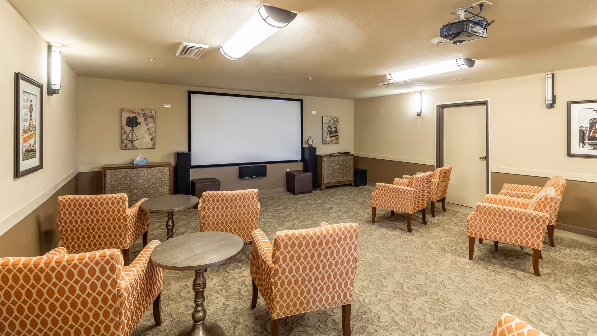 A cozy media room with a large projector screen on the far wall, surrounded by speakers and cabinets. The room has patterned carpet and beige walls with framed artwork. There are eight orange patterned armchairs arranged in pairs, each pair separated by small round tables. Ceiling lights and wall sconces provide warm lighting.