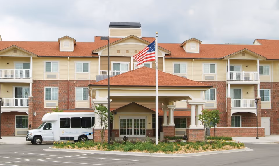 Front entrance of the Shawnee Hills senior living building with a porte-cochere, American flag, and a shuttle van parked nearby.