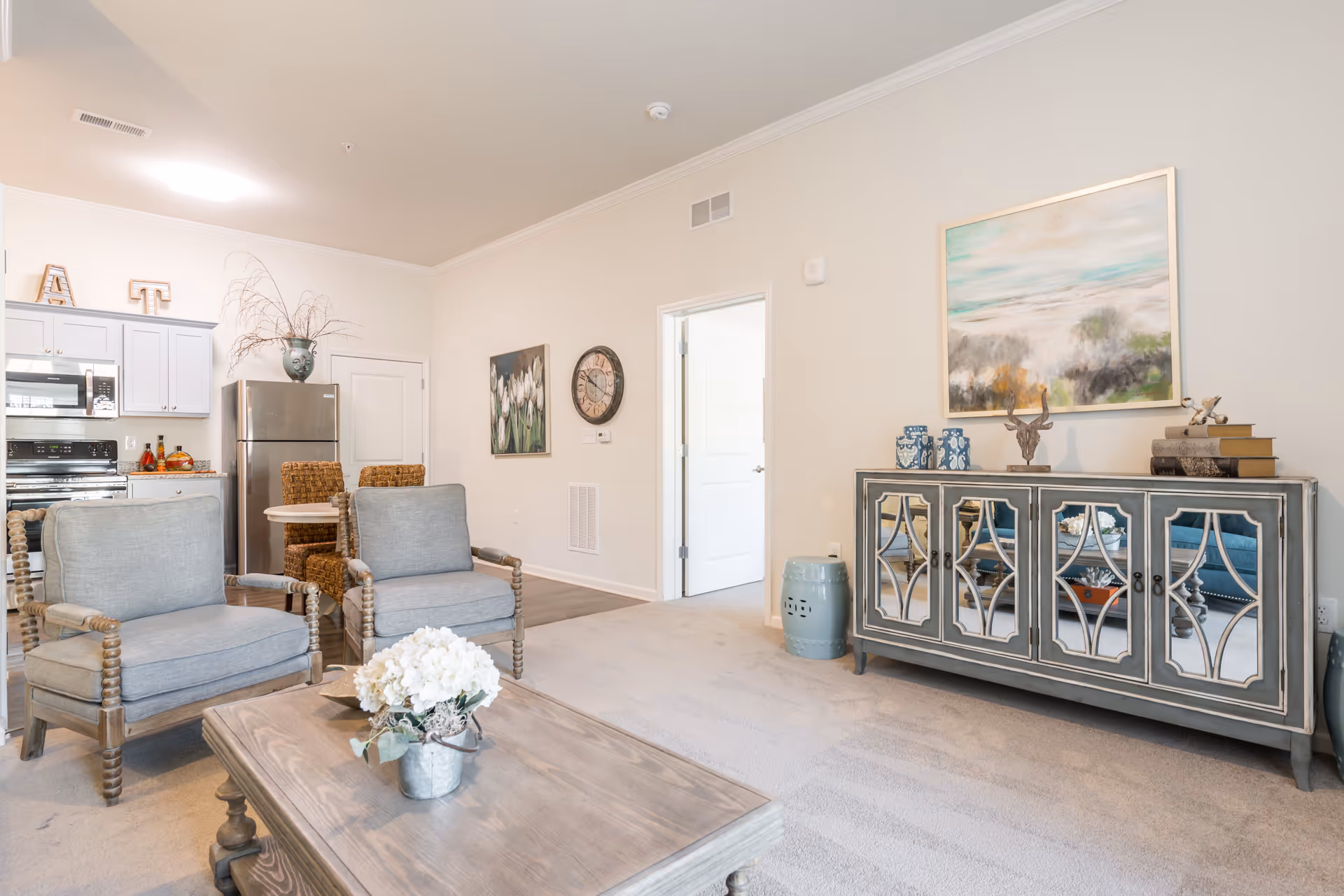 A bright and airy living room area with two gray cushioned armchairs and a wooden coffee table with a small flower arrangement. In the background, there is a kitchen area with white cabinets, a stainless steel refrigerator, and a small dining table with woven chairs. A decorative sideboard with mirrored doors and various decorative items is against the wall, along with a large framed painting above it. The walls are light-colored, and the room has a cozy, welcoming atmosphere.