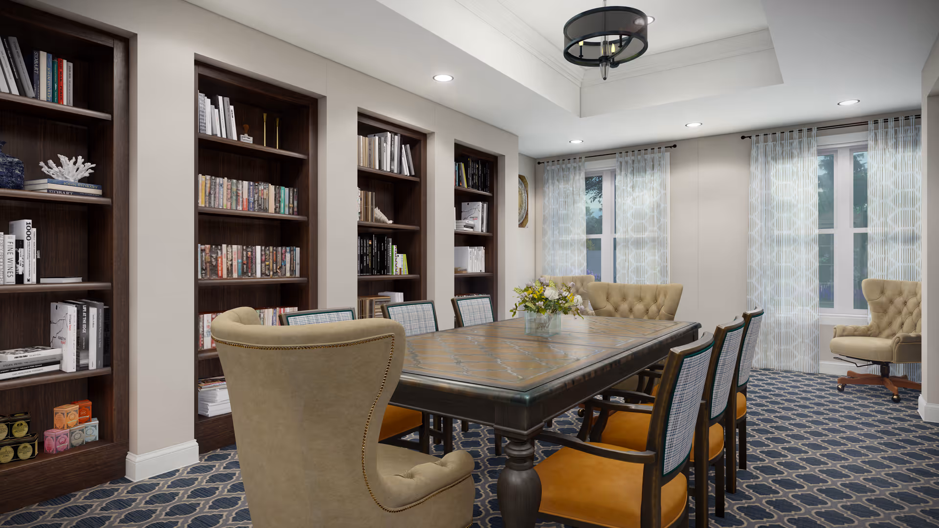 A well-lit room with a large wooden table surrounded by eight chairs, including two upholstered armchairs at the ends and six wooden chairs with orange cushions. The room features built-in dark wood bookshelves filled with books and decorative items along one wall. Two windows with sheer patterned curtains allow natural light to enter. The carpet has a blue and white geometric pattern, and a floral arrangement is placed in the center of the table.