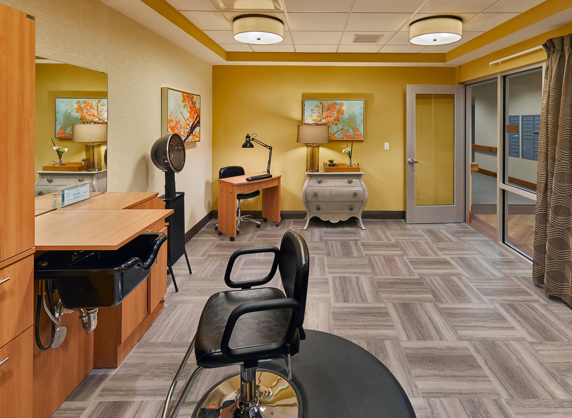 A well-lit salon area in a nursing home with a black salon chair in front of a wooden counter with a sink. There is a hair dryer on a stand, a small desk with a lamp and chair, a decorative chest with a lamp and flowers, and a painting of orange flowers on the wall. The room has a yellow accent wall, tiled flooring, and a glass door leading to a hallway.