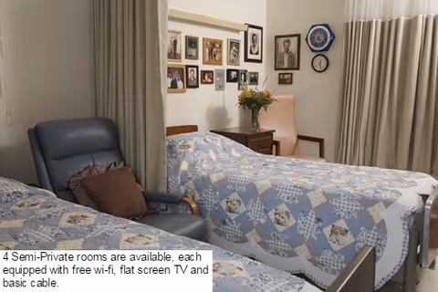 A semi-private nursing home room with two beds covered in blue and white patterned quilts. Between the beds is a wooden nightstand with a vase of flowers. A blue recliner chair with a brown pillow is next to one bed. The walls have several framed photographs and a clock, and beige curtains cover the windows.