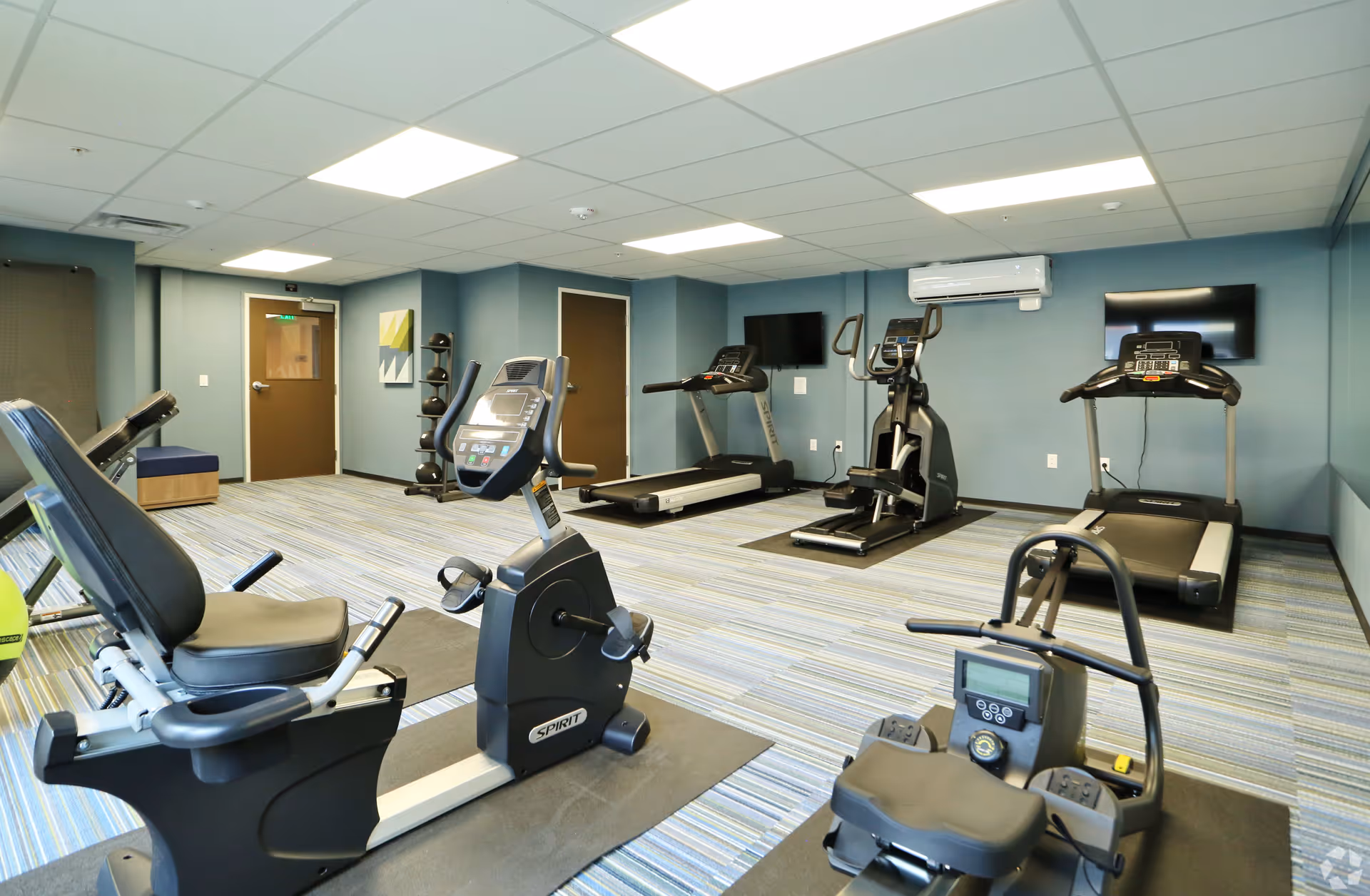 A fitness room with various exercise equipment including stationary bikes, treadmills, an elliptical machine, and a rowing machine. The room has blue walls, a striped carpet, two wall-mounted TVs, and bright ceiling lights.