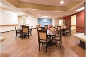 A spacious dining room with several square wooden tables and cushioned chairs arranged neatly on a wood floor. The walls are painted in neutral tones with some accent colors, and there is a flat-screen TV mounted on one wall. The room is well-lit with recessed ceiling lights.