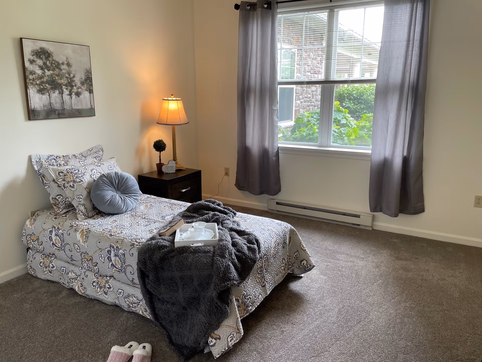 A neatly made single bed with floral patterned bedding and a round gray pillow in a bedroom. A dark wooden nightstand with a small lamp, a small potted plant, and a decorative sign is beside the bed. A window with gray curtains lets in natural light, showing greenery outside. A pair of slippers is on the carpeted floor near the bed.
