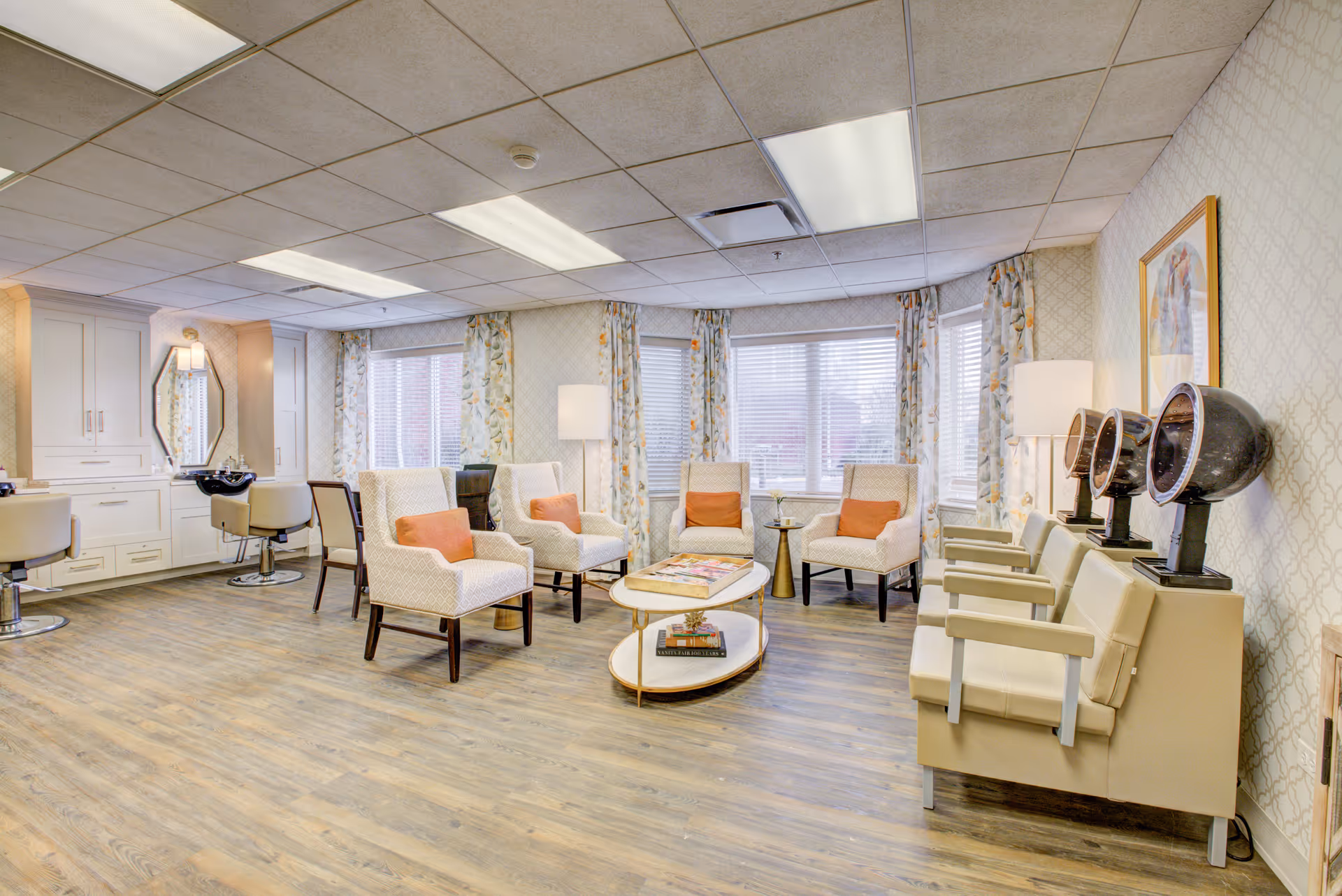 A bright and spacious salon area in a senior living facility with several beige armchairs featuring orange cushions arranged around a white oval coffee table. On the right side, there are three vintage-style hair dryers mounted on beige chairs. The room has large windows with floral curtains, wood flooring, and soft lighting from ceiling fixtures and floor lamps.