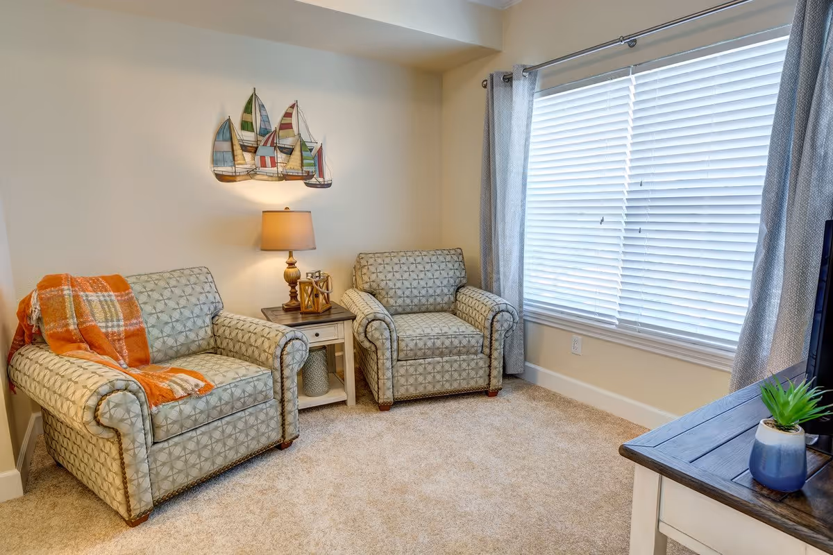 A cozy living room area with two patterned armchairs, one draped with an orange plaid blanket, a small wooden side table with a lamp and decorative items, a large window with closed blinds and gray curtains, a wooden TV stand with a small potted plant, and a wall decoration featuring colorful sailboats.