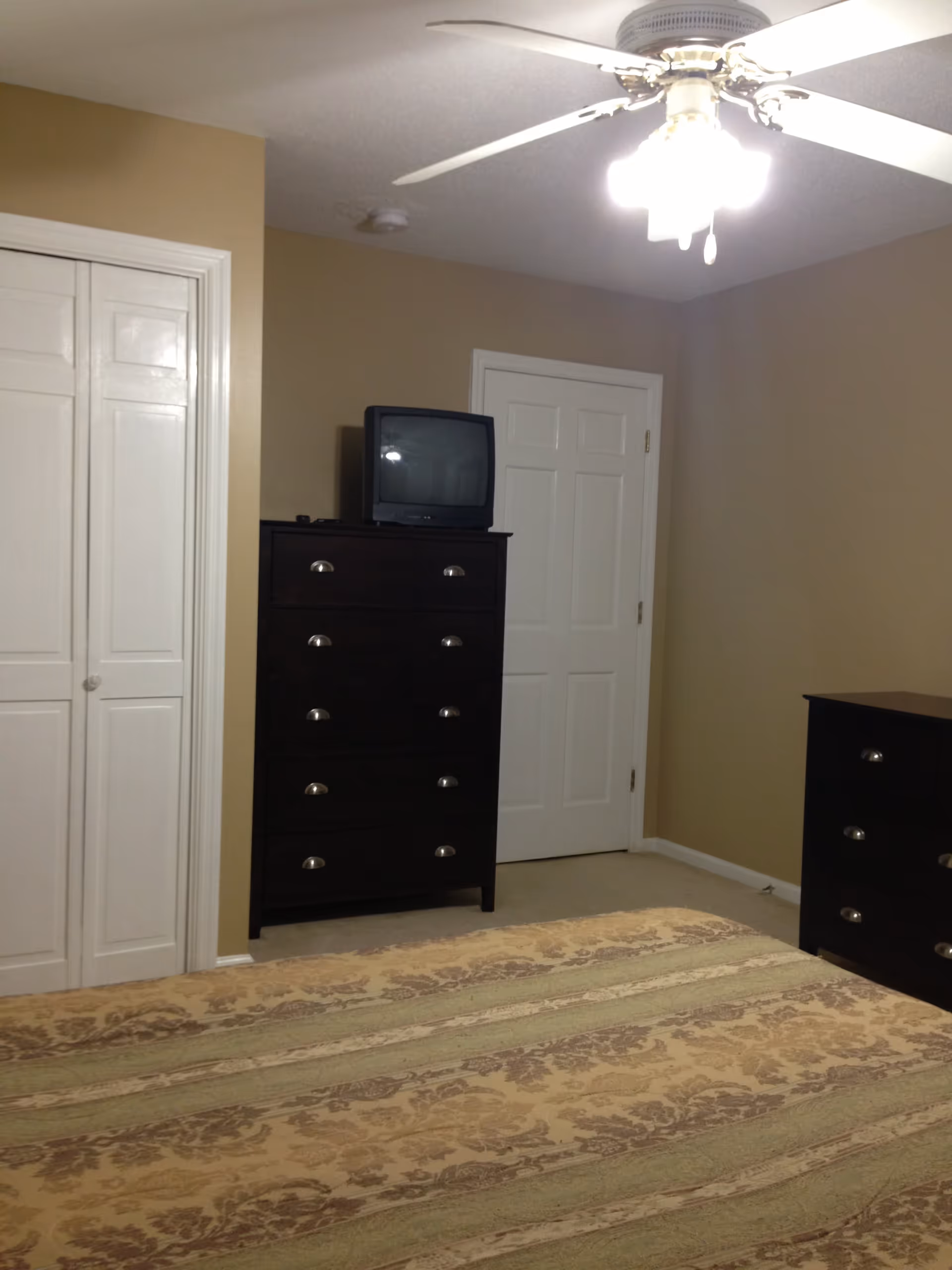 A bedroom with beige walls and carpeted floor. There is a bed with a patterned bedspread in the foreground. Against the wall, there is a dark wooden dresser with a small CRT television on top. To the right, there is another dark wooden dresser. A white door and a white closet door are visible on the back wall. A ceiling fan with lights is on the ceiling.